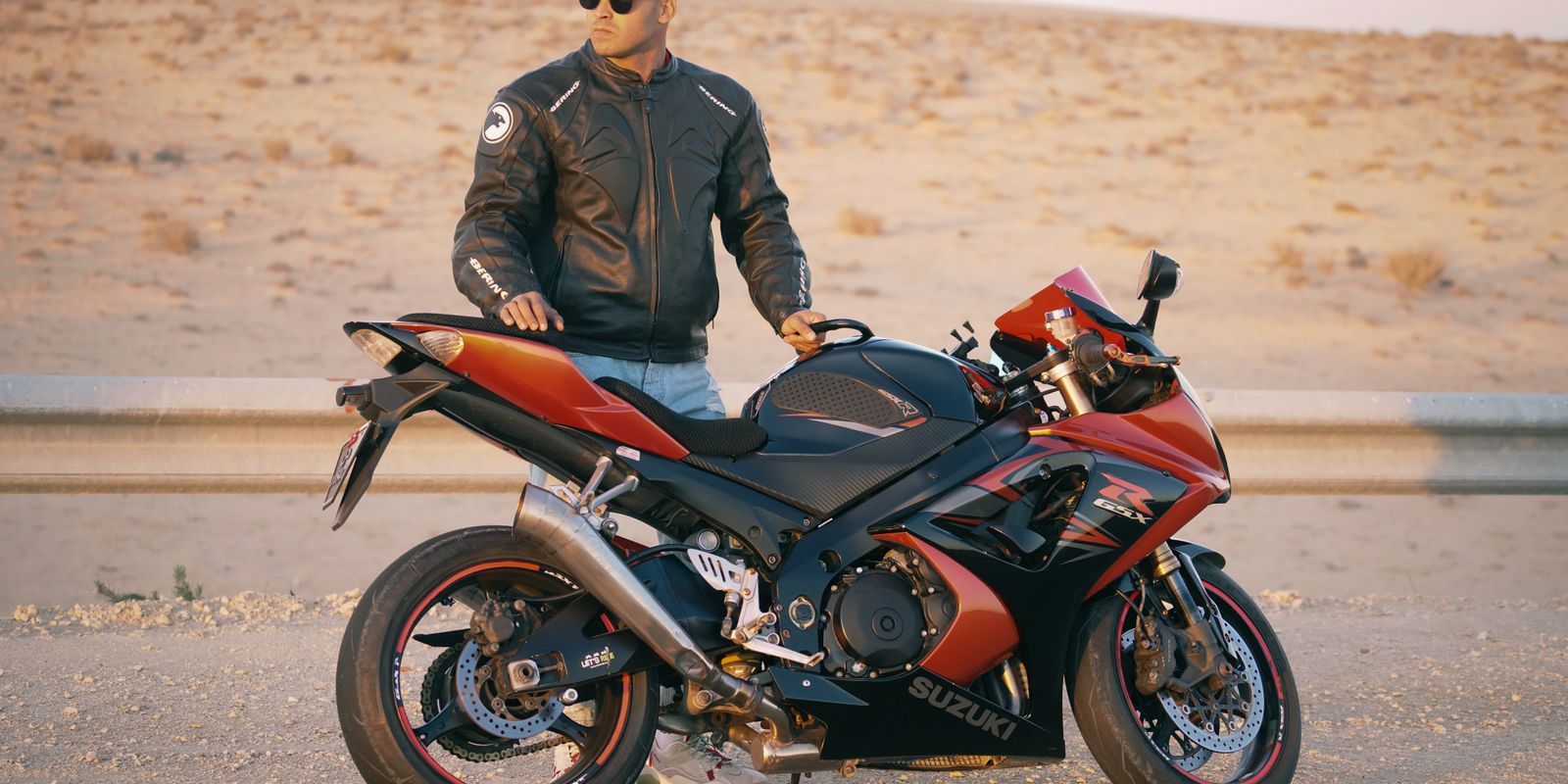 Buneri.fr | Bomber, motard ou racer : quel style de veste en cuir vous correspond ? Homme en veste en cuir et casquette posant avec une moto sportive sur une route désertique