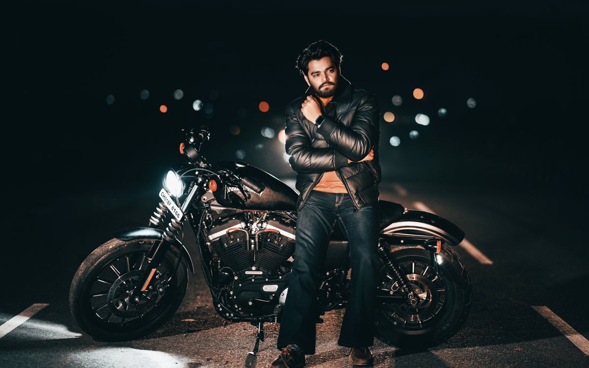 Buneri.fr | Bomber, motard ou racer : quel style de veste en cuir vous correspond ? Homme en veste en cuir posant avec une moto de nuit, lumières de la ville en arrière plan