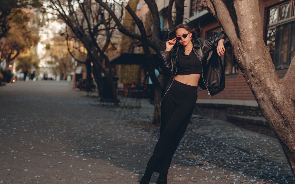 Femme élégante en tenue noire posant dans un décor urbain automnal avec des feuilles au sol
