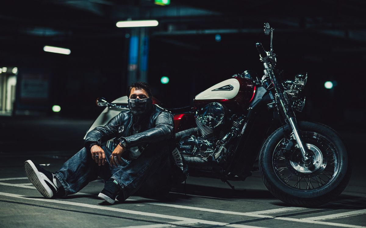 Motard en veste en cuir agenouillé près d'une moto dans un garage faiblement éclairé