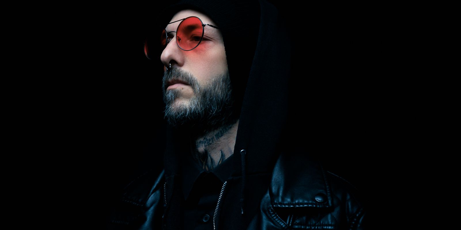 Buneri.fr | Veste en Cuir sur un Hoodie : Réussir le Streetwear Portrait mélancolique d'un homme en veste de cuir noire et lunettes teintées rouges sur fond sombre