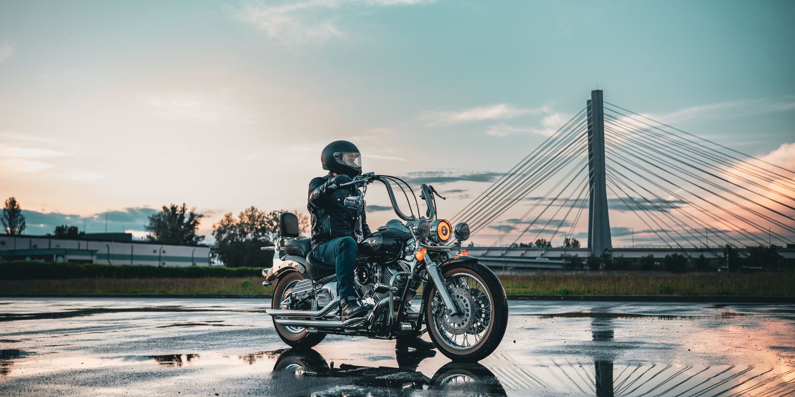 Motard en tenue de cuir sur une route mouillée avec un pont en arrière-plan au coucher du soleil