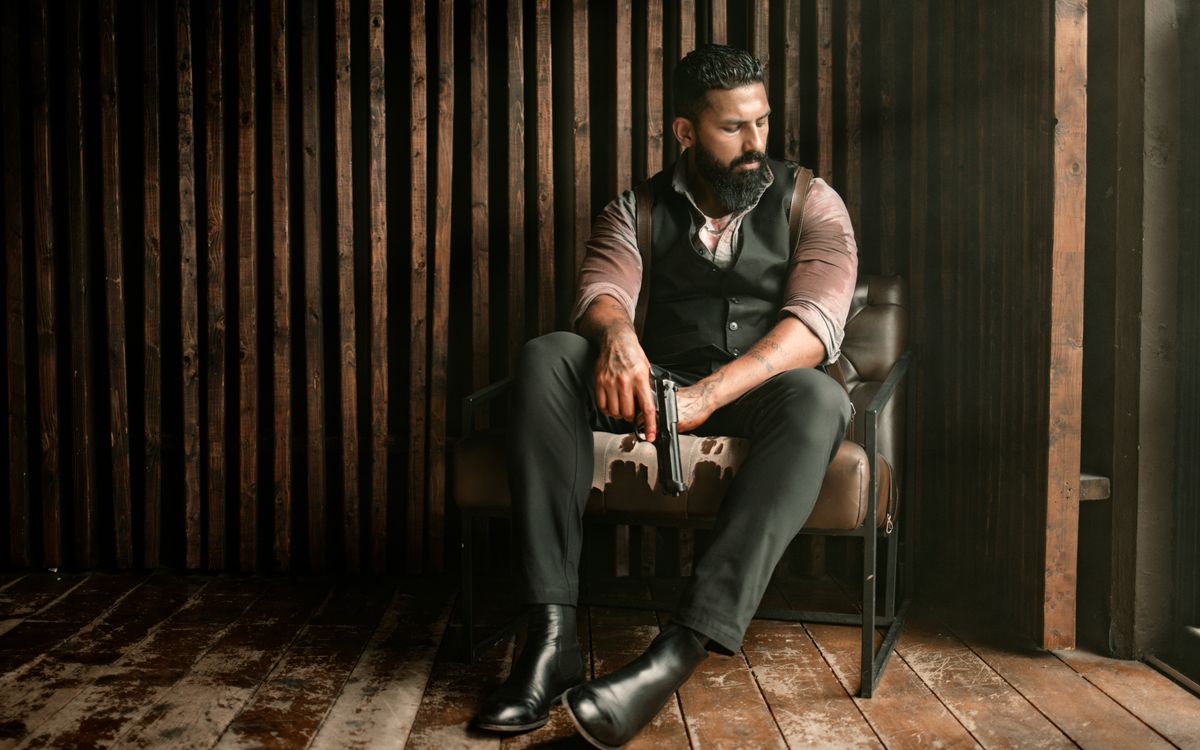 Homme barbu portant un gilet, assis à l'intérieur devant un décor rustique