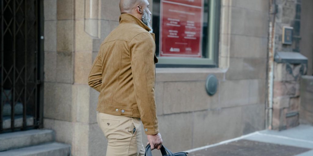 Un homme barbu marche dans une rue en portant une mallette, vêtu d'une veste beige