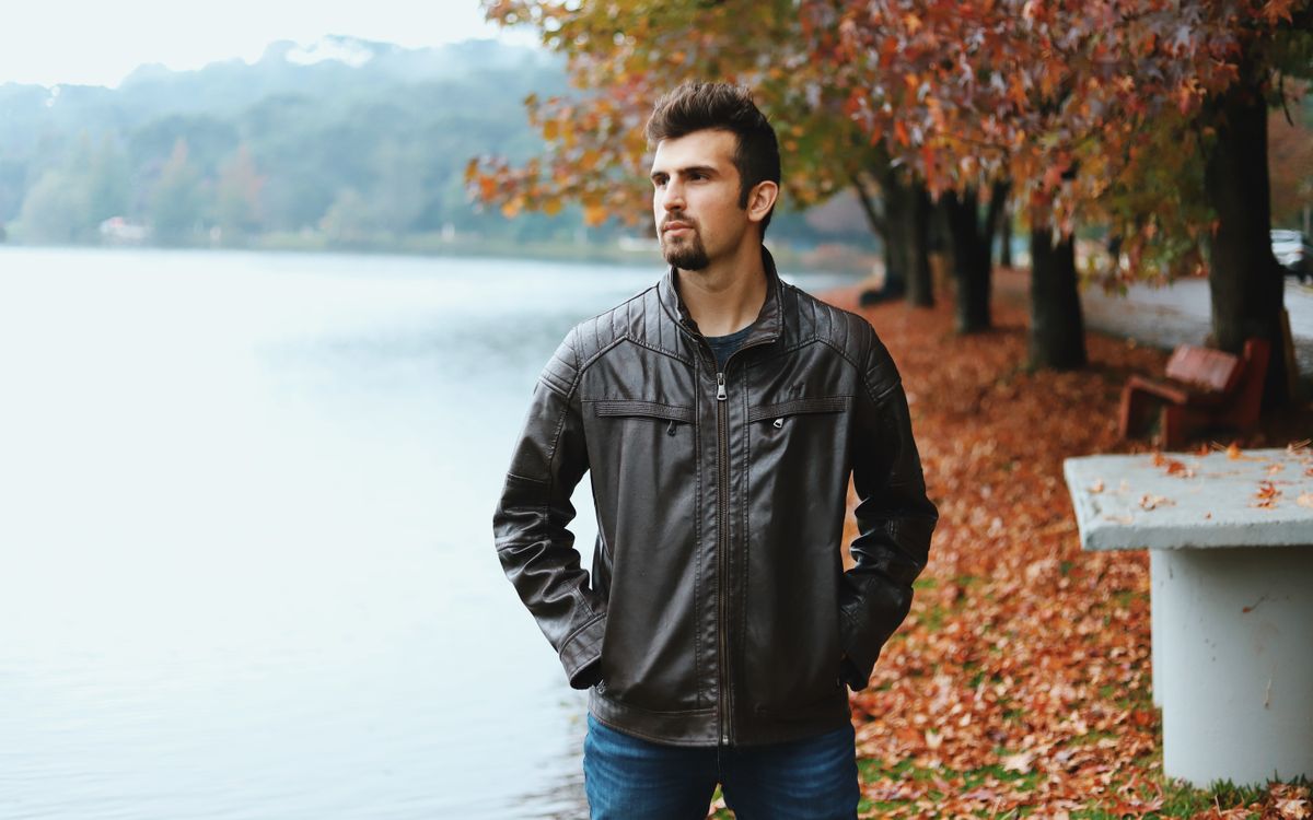 Un homme barbu en veste de cuir marron au bord d'un lac en automne