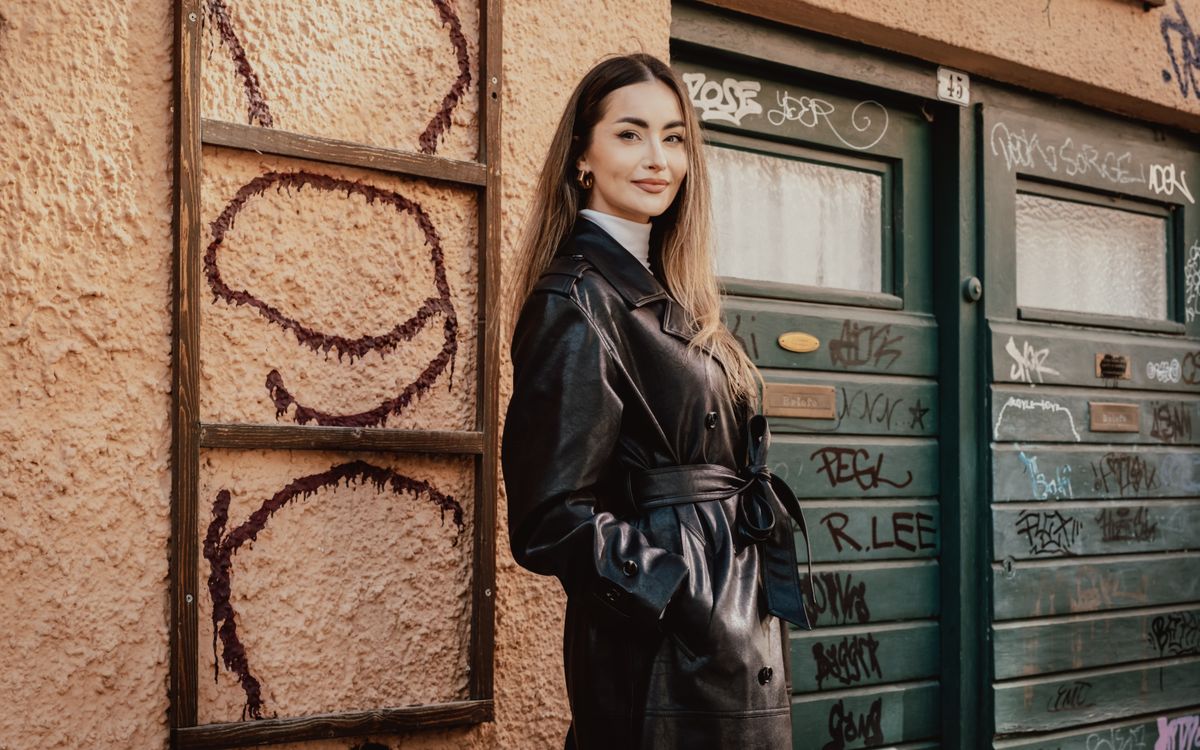 Une femme pose avec assurance dans un long manteau en cuir noir devant un mur urbain