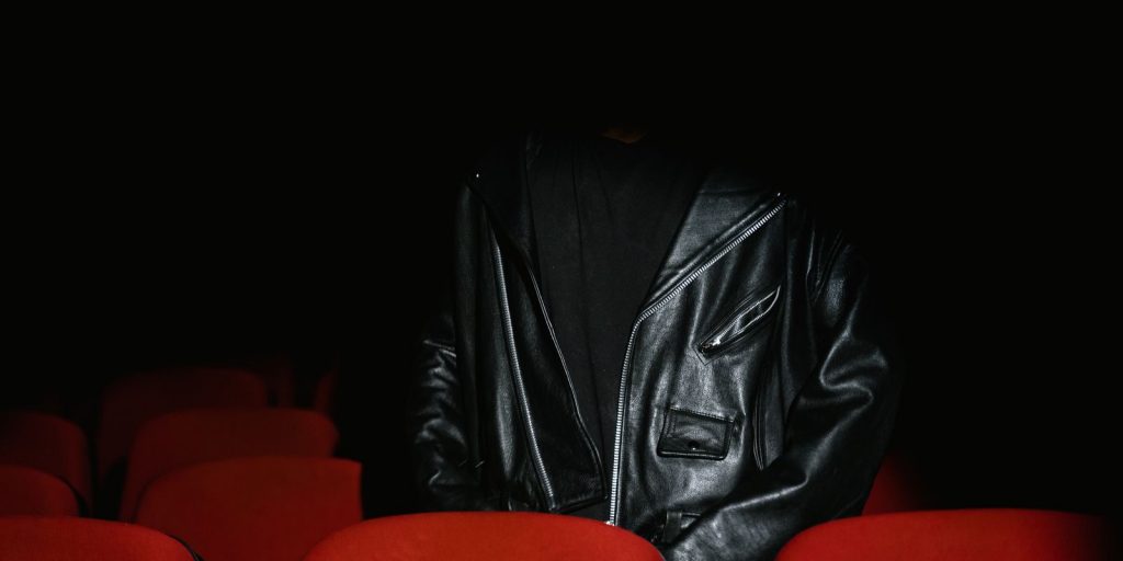 Une personne en veste de cuir noir assise dans une salle de cinéma tamisée aux sièges rouges