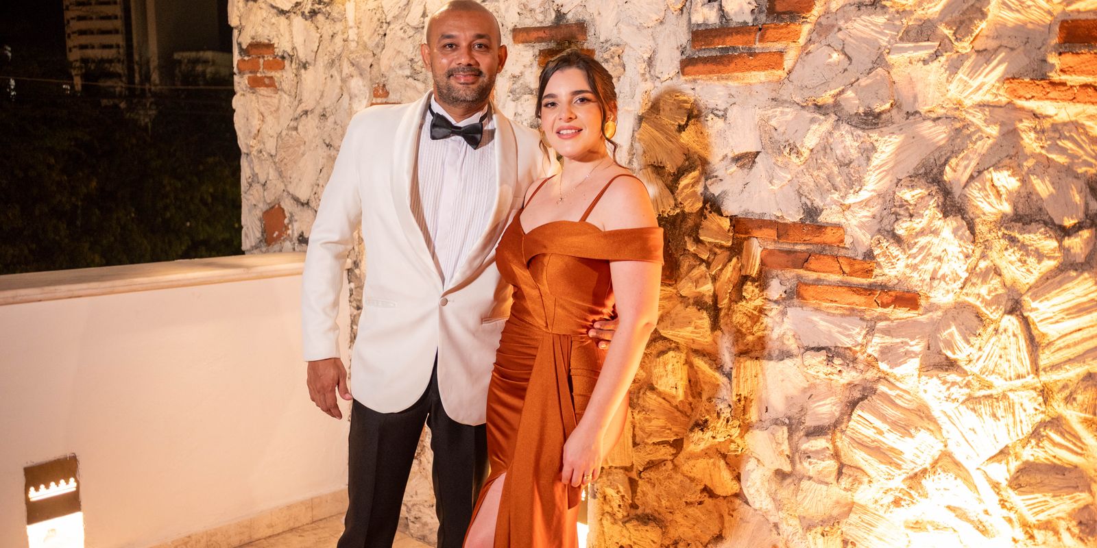 Couple élégant en tenue de soirée posant ensemble devant un mur de pierre illuminé